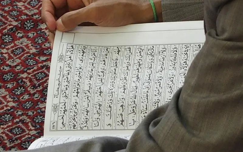 Homme assis sur un tapis de prière lisant une page du Coran ouverte, illustrant l’apprentissage et la récitation au quotidien