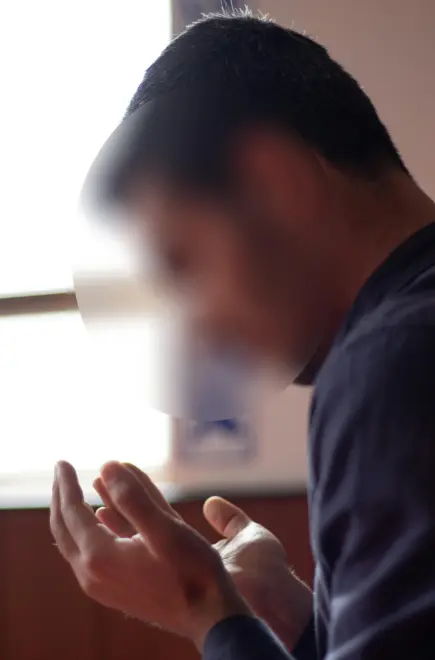 Man praying with raised hands near a window, bathed in natural light, expressing serenity and spiritual reflection.